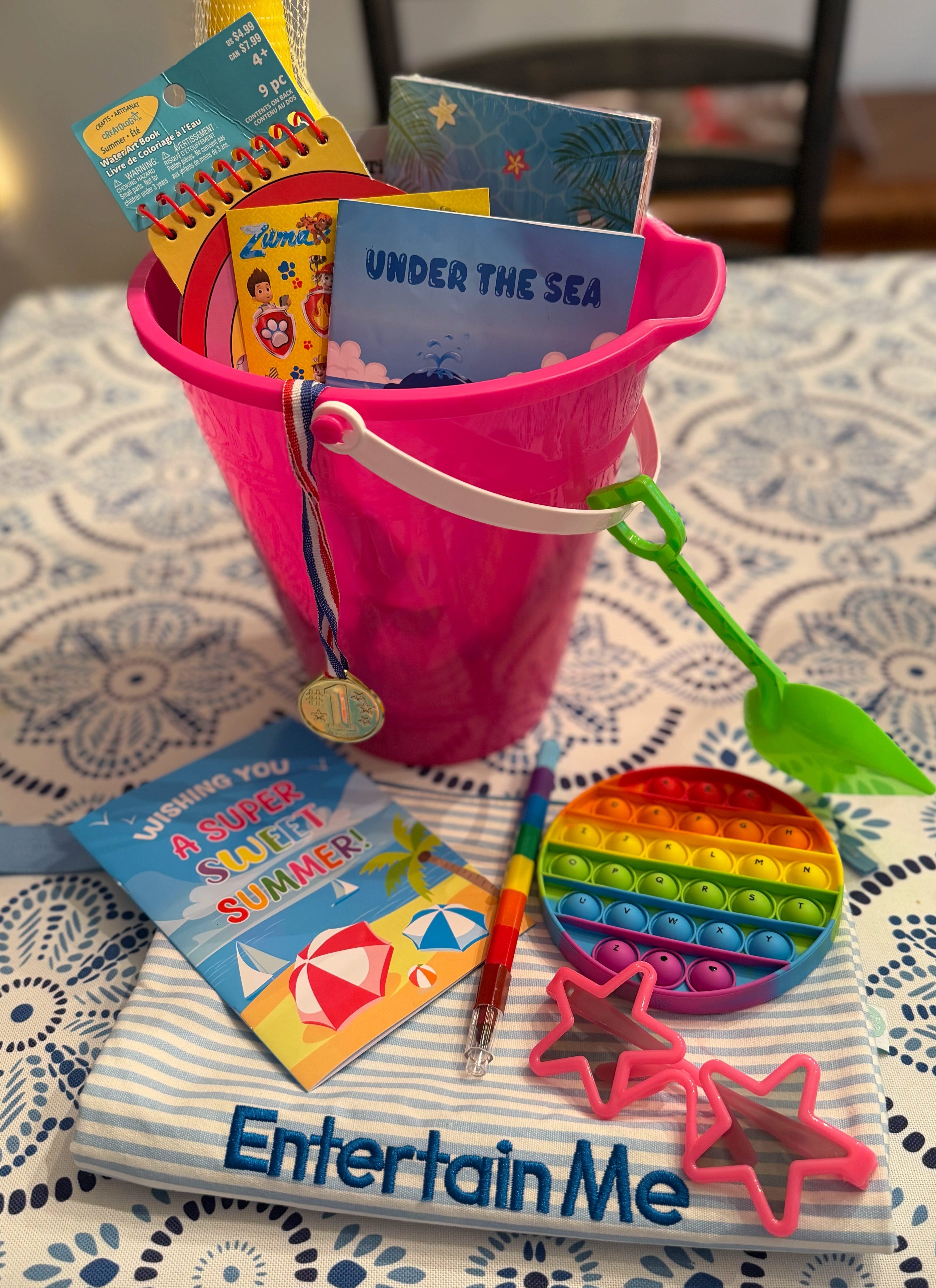 Small coloring book, pop-it, sunglasses, and pen arranged with an Entertain Me pouch for pool break activities.