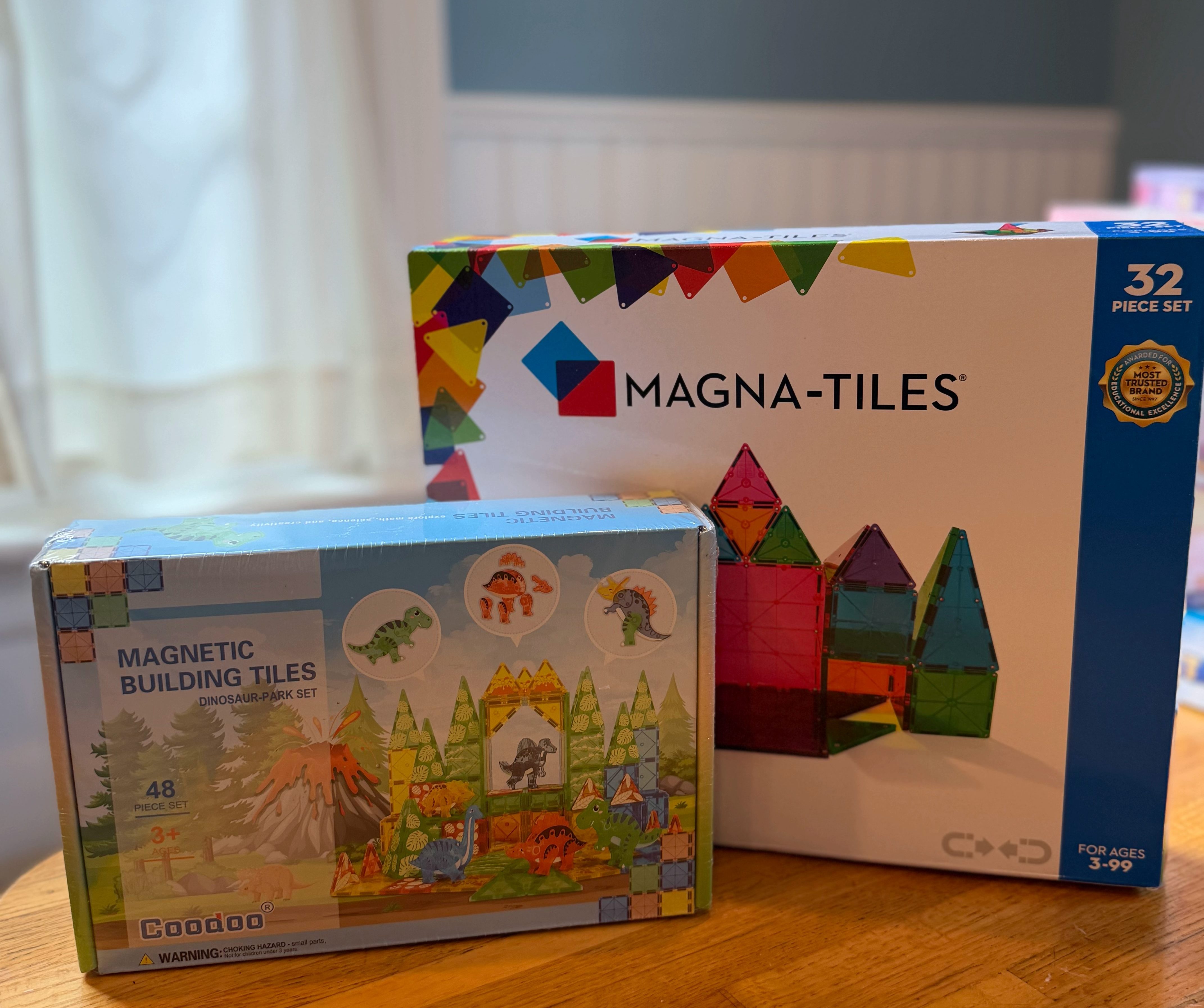 Two boxes of magnetic building tiles, including a dinosaur-themed set and a Magna-Tiles set, arranged on a wooden table.