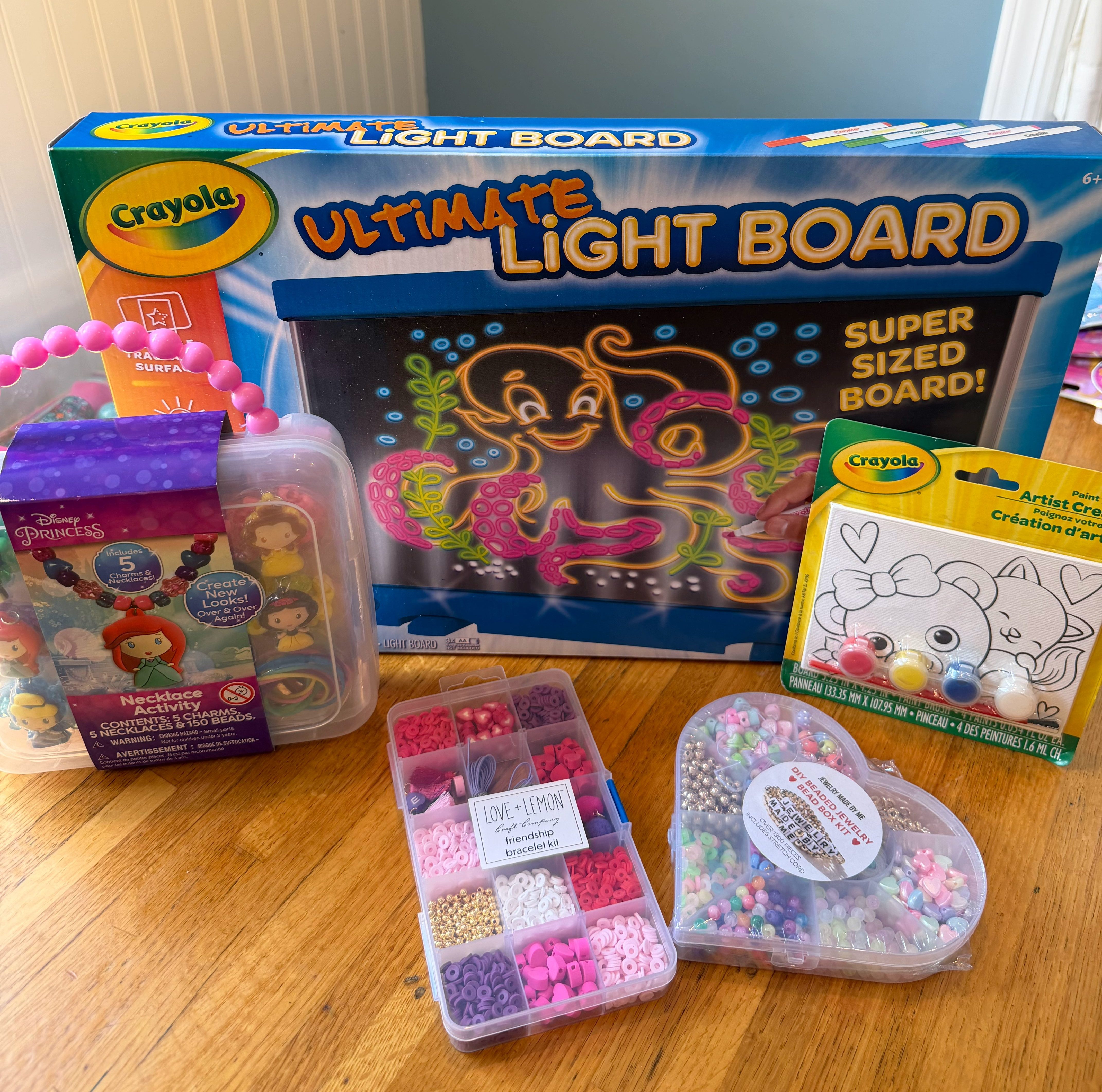 Craft-themed birthday gifts including a Crayola light board, Disney princess necklace kit, bead kits, painting activity, and small craft supplies arranged on a wooden table.